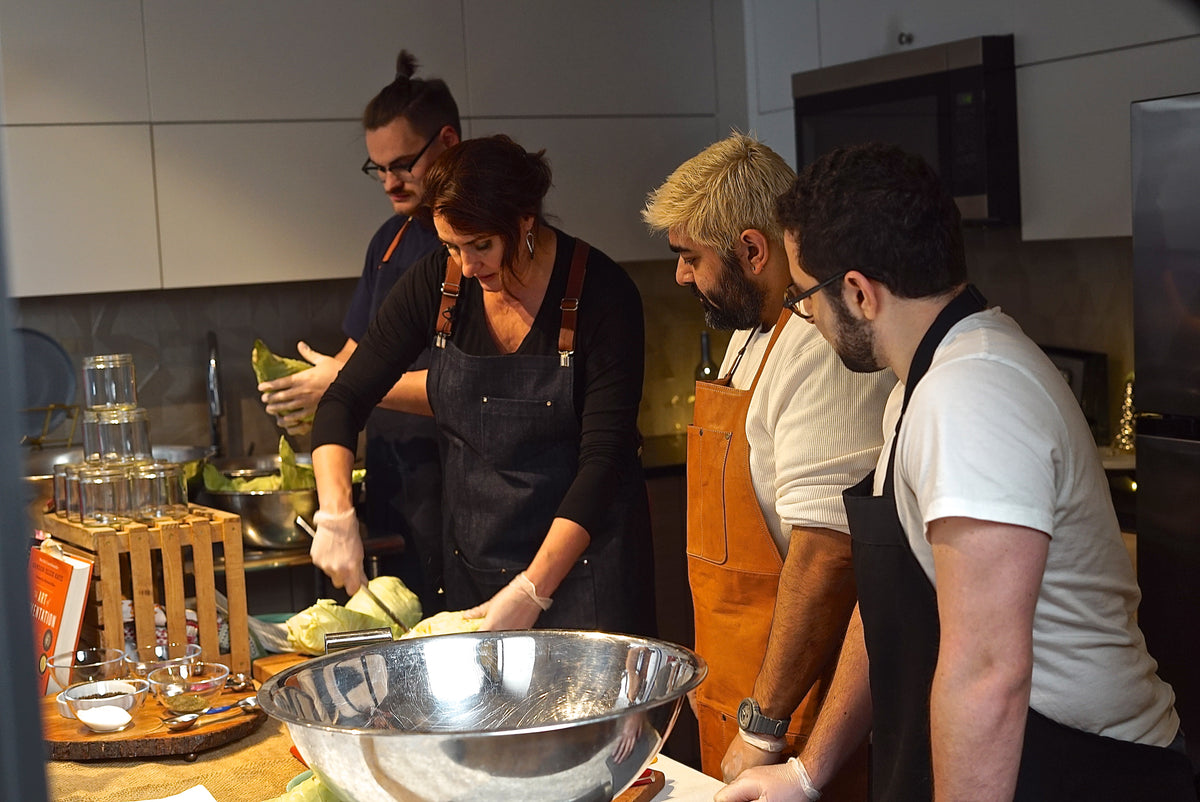 Four people in a modern kitchen preparing food, wearing aprons and gloves around a large stainless steel mixing bowl on a wooden counter.
